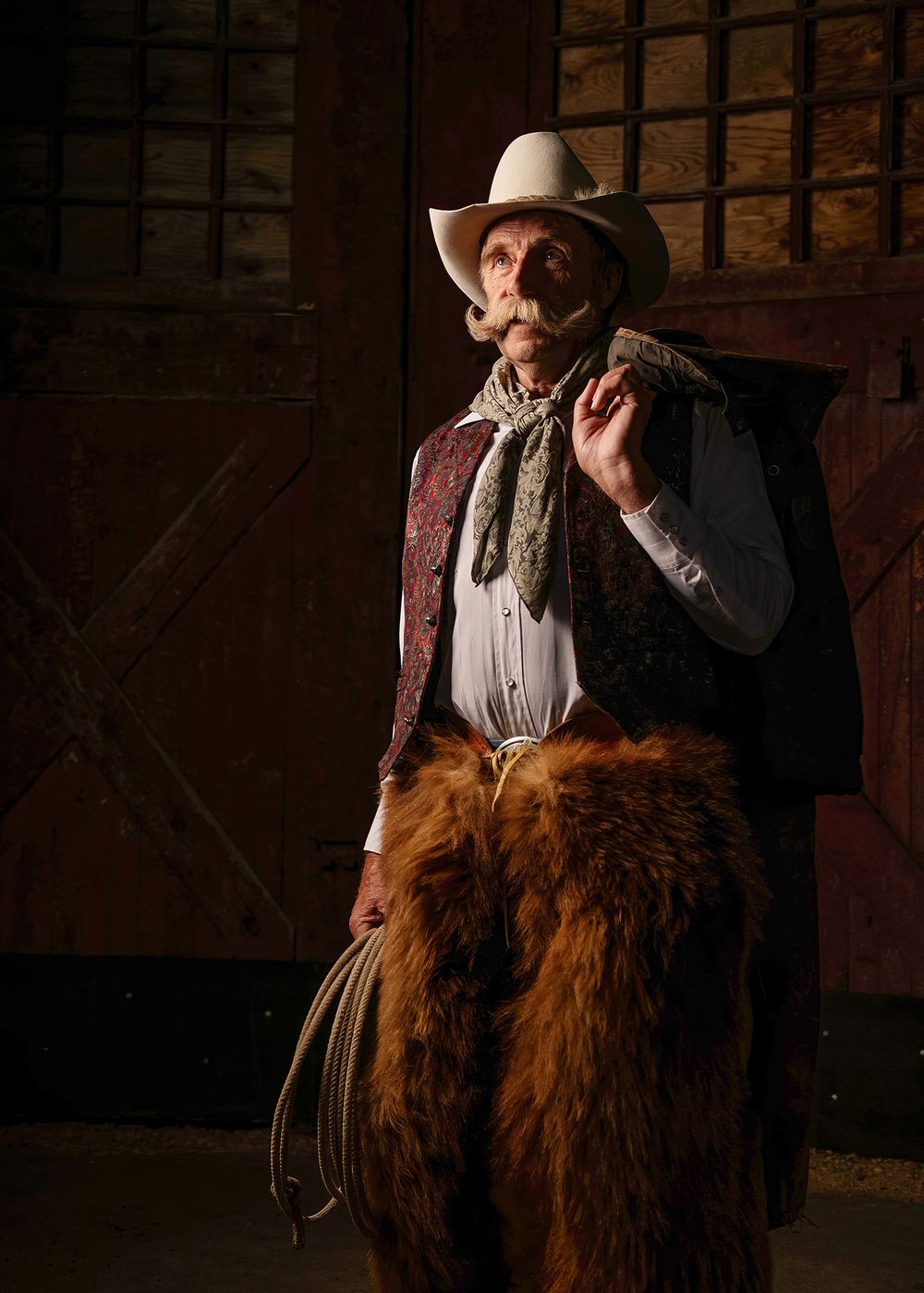 Older cowboy in white hat, maroon vest, and fur chaps  hohlding rope and coat, looking upward - Okanagan portrait by Tania Veitch.