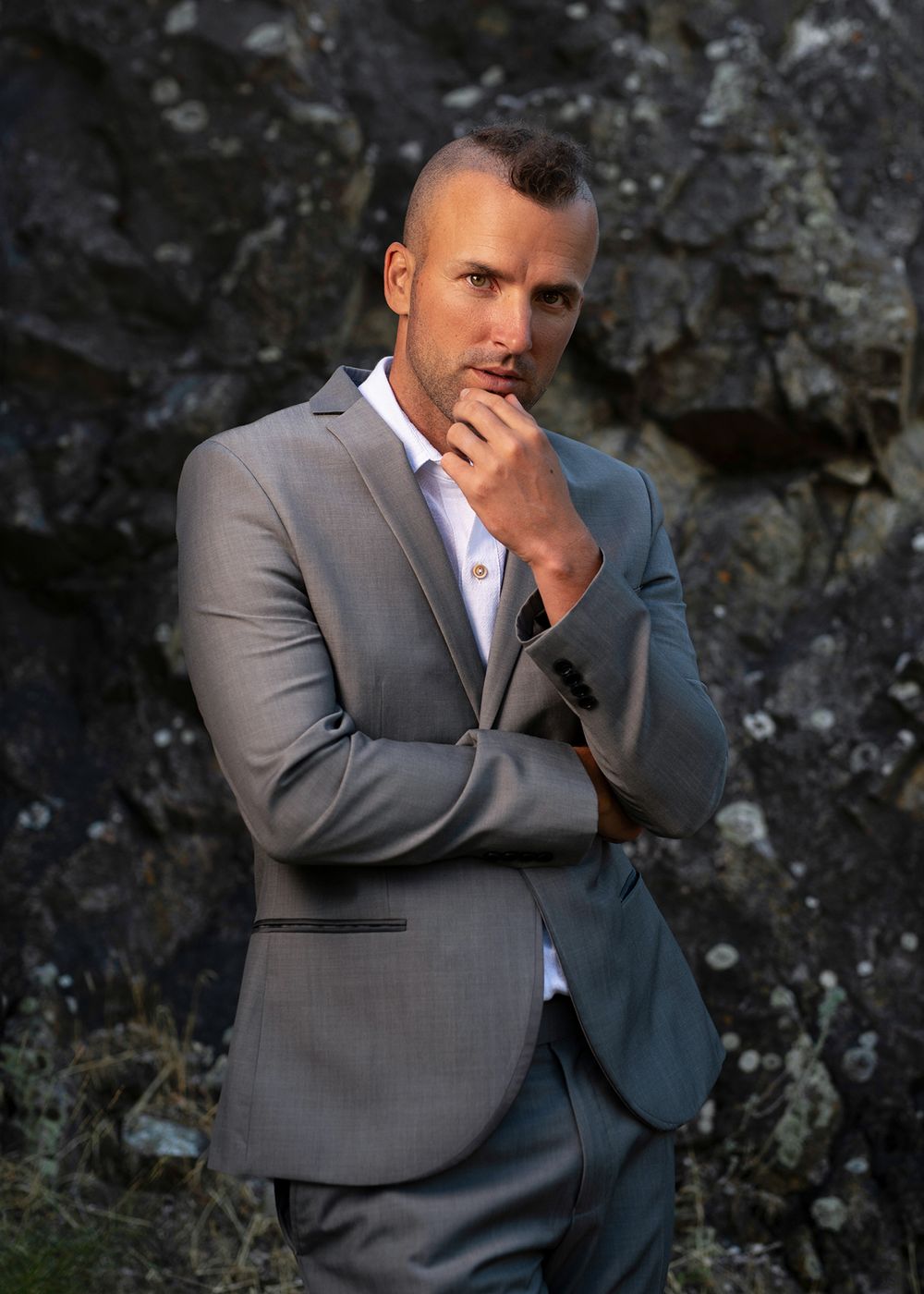 Young man in grey suit and white shirt posing with hand on chin before rock wall  - Okanagan portrait by Tania Veitch.
