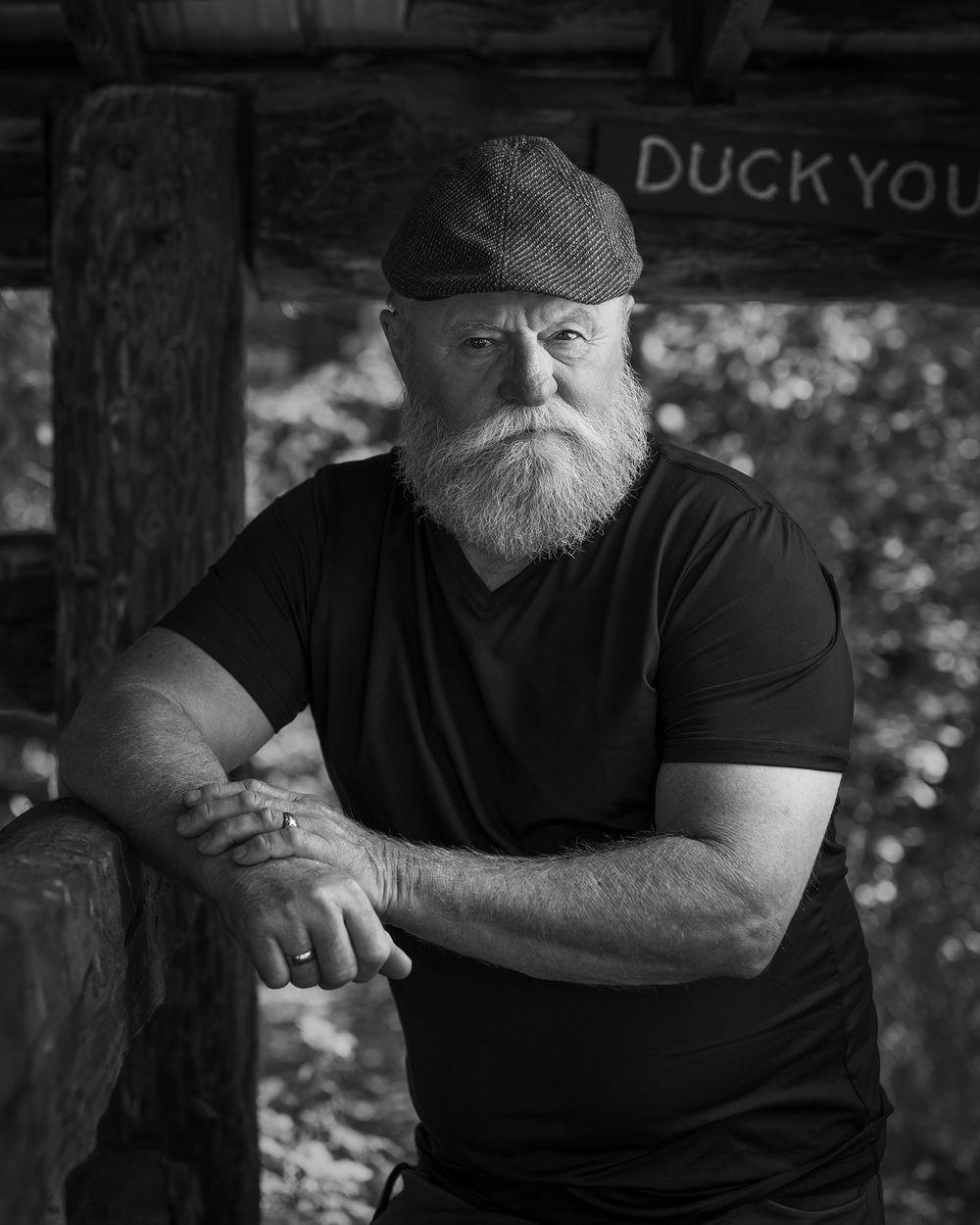 Black and white environmental portrait of bearded man in flat cap outdoors.