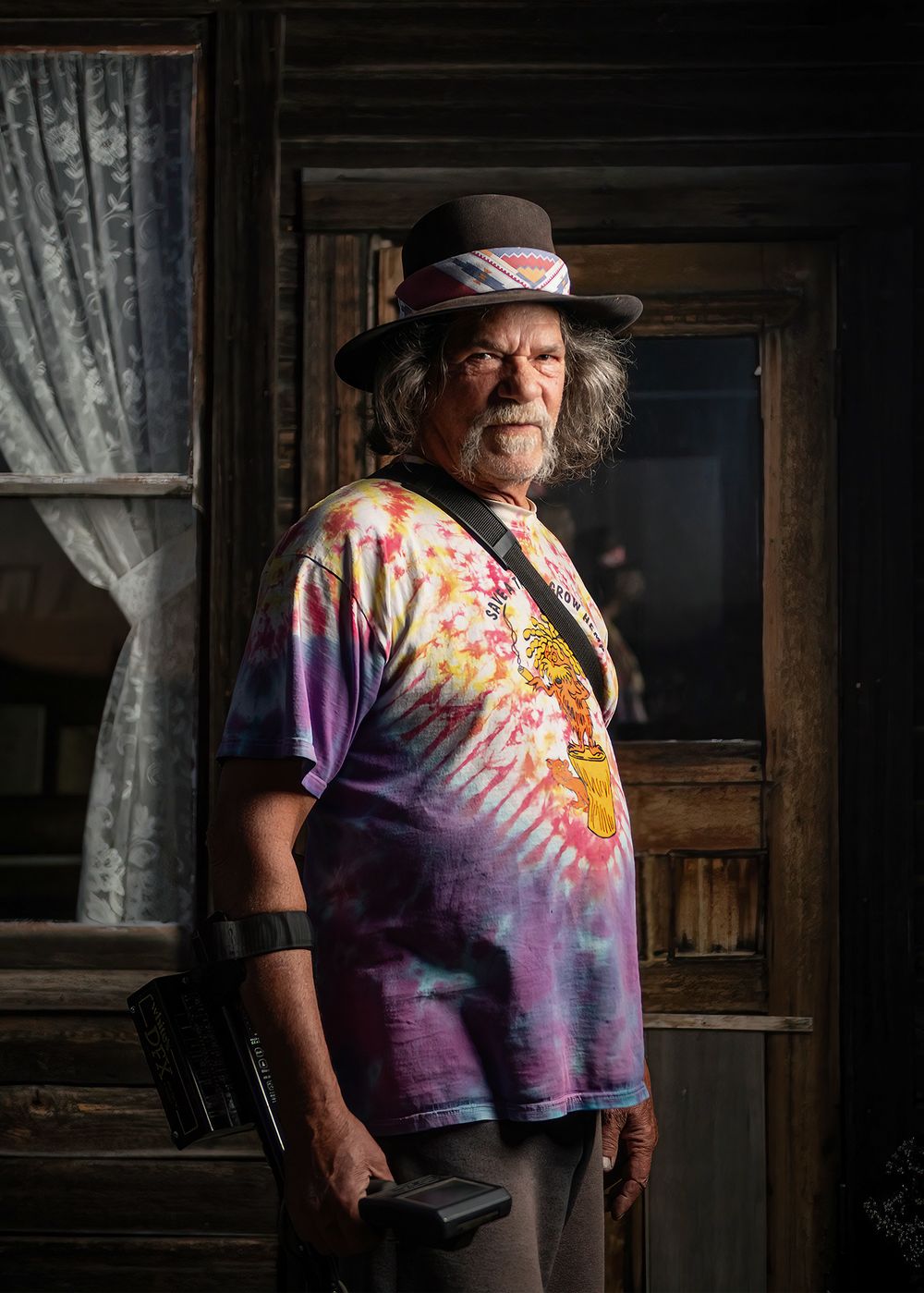 Older man with beard, black hat, and tie-dye shirt holding metal detector before weathered house - Okanagan portrait by Tania Veitch.
