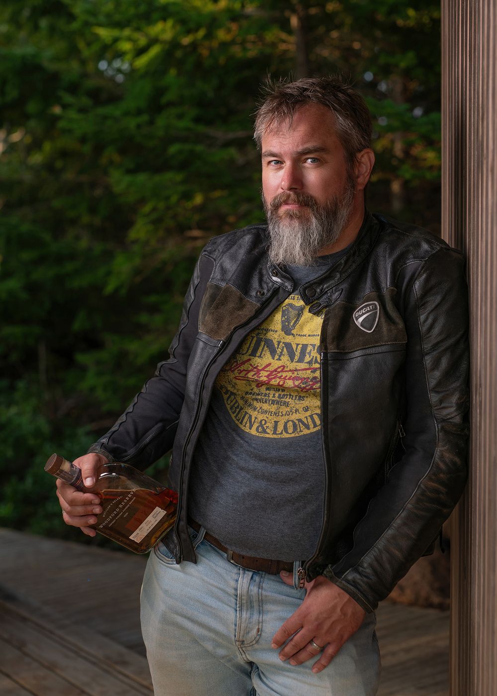 Middle-aged man with beard in black leather jacket leaning against wall holding whiskey bottle - Okanagan portrait by Tania Veitch.