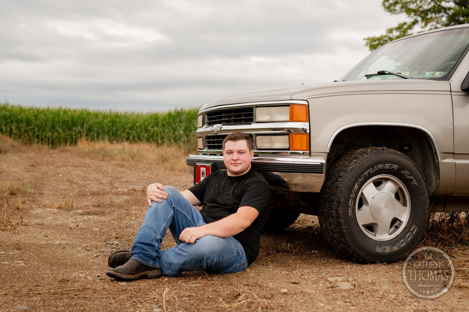 A person sits relaxed against a silver Chevrolet truck on a dirt road by a cornfield.