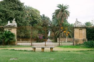 Ornate iron gates and stone pillars leading to a lush garden with palm trees.