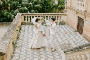 Wedding couple stands on ornate tiled terrace with palm trees and classic architecture.