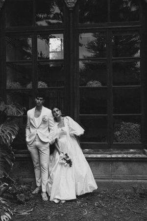 A black and white wedding portrait of a couple posing against large windowpanes.