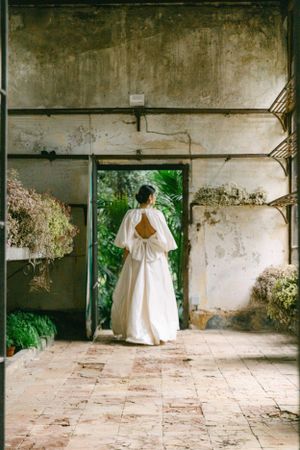 A bride in a white dress with open back detail stands in doorway of rustic building.