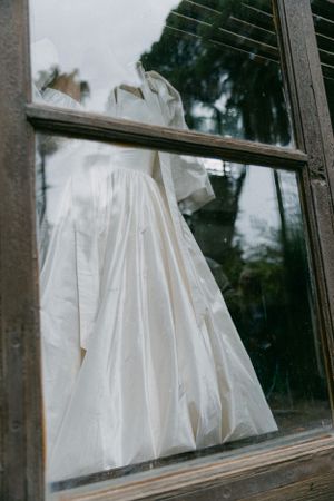 A white wedding dress photographed through a window pane with reflection of trees.