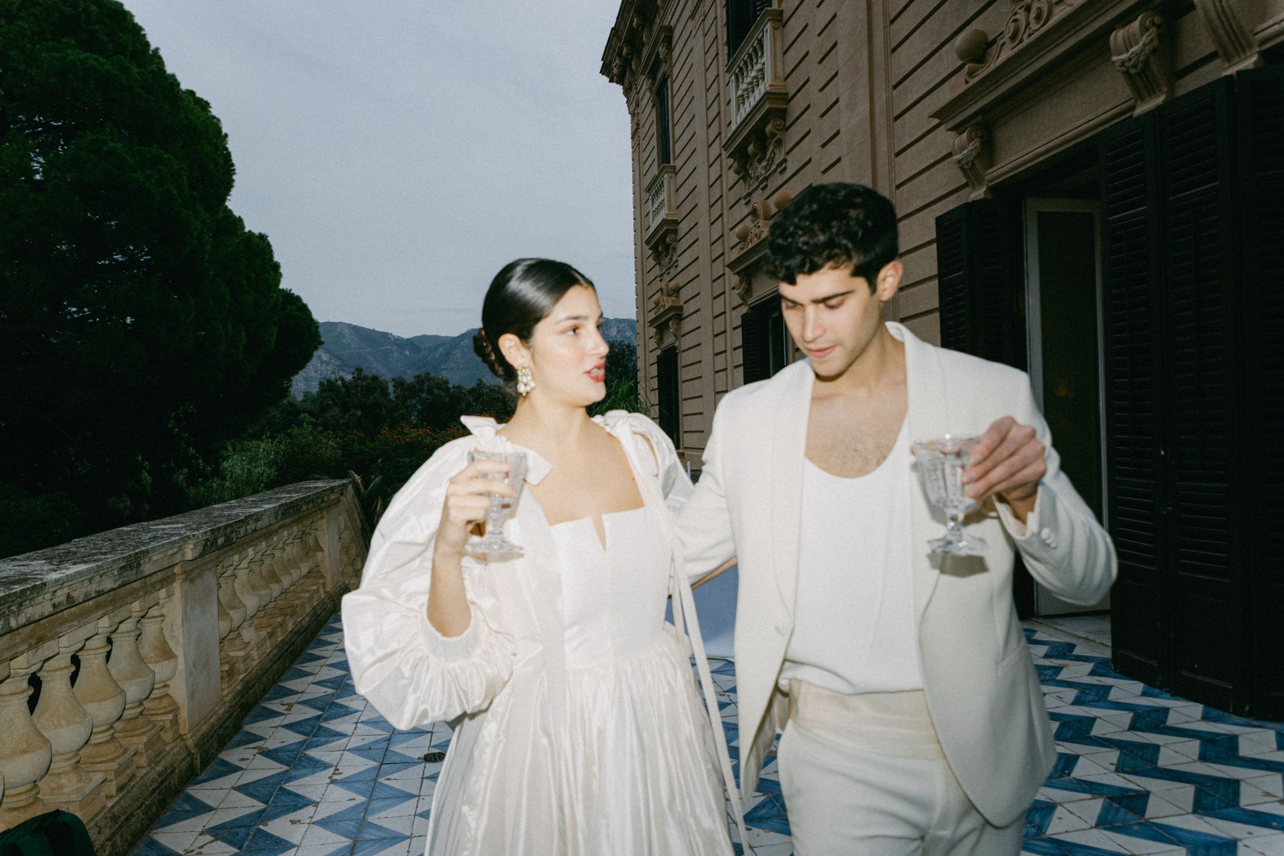 A candid moment of a couple in white attire sharing drinks on a checkered floor terrace.