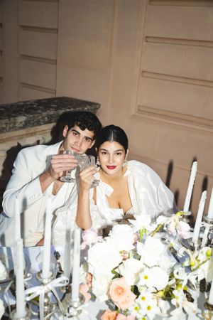 A romantic dinner scene with candles and floral arrangements as a couple toasts with glasses.