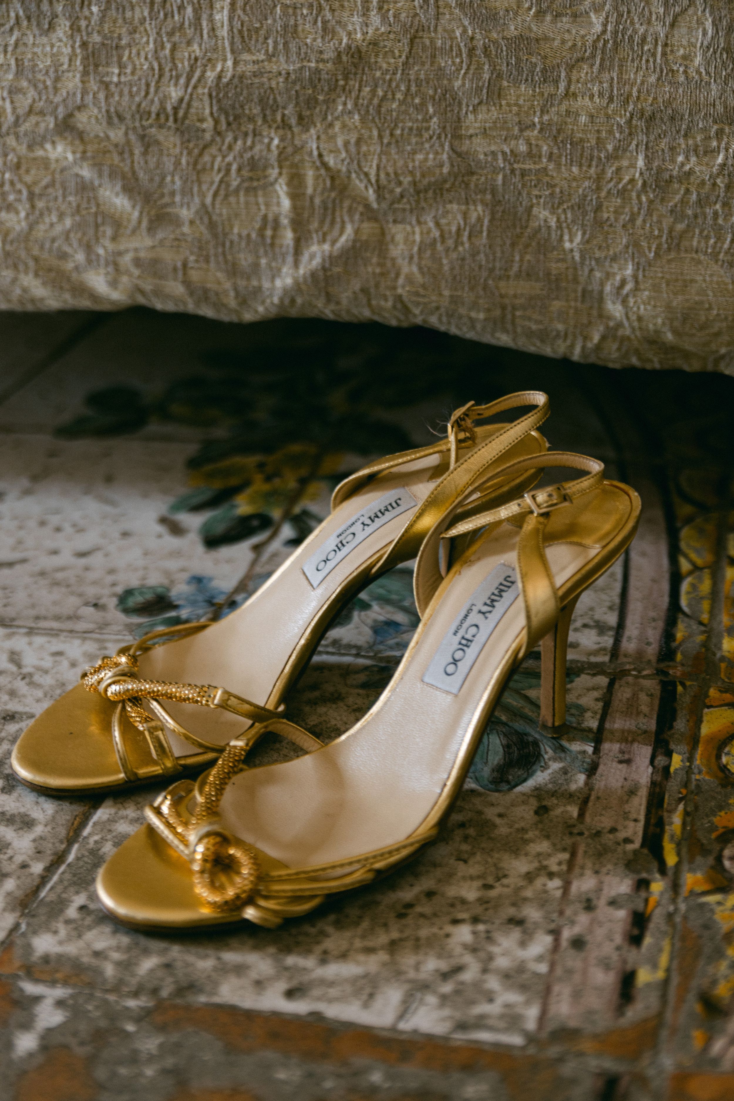 Gold Jimmy Choo sandals with bow detail photographed on vintage surface.