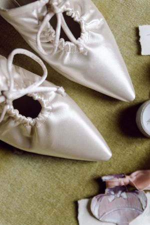 White satin wedding shoes with pointed toes and gathered details rest on a green surface.