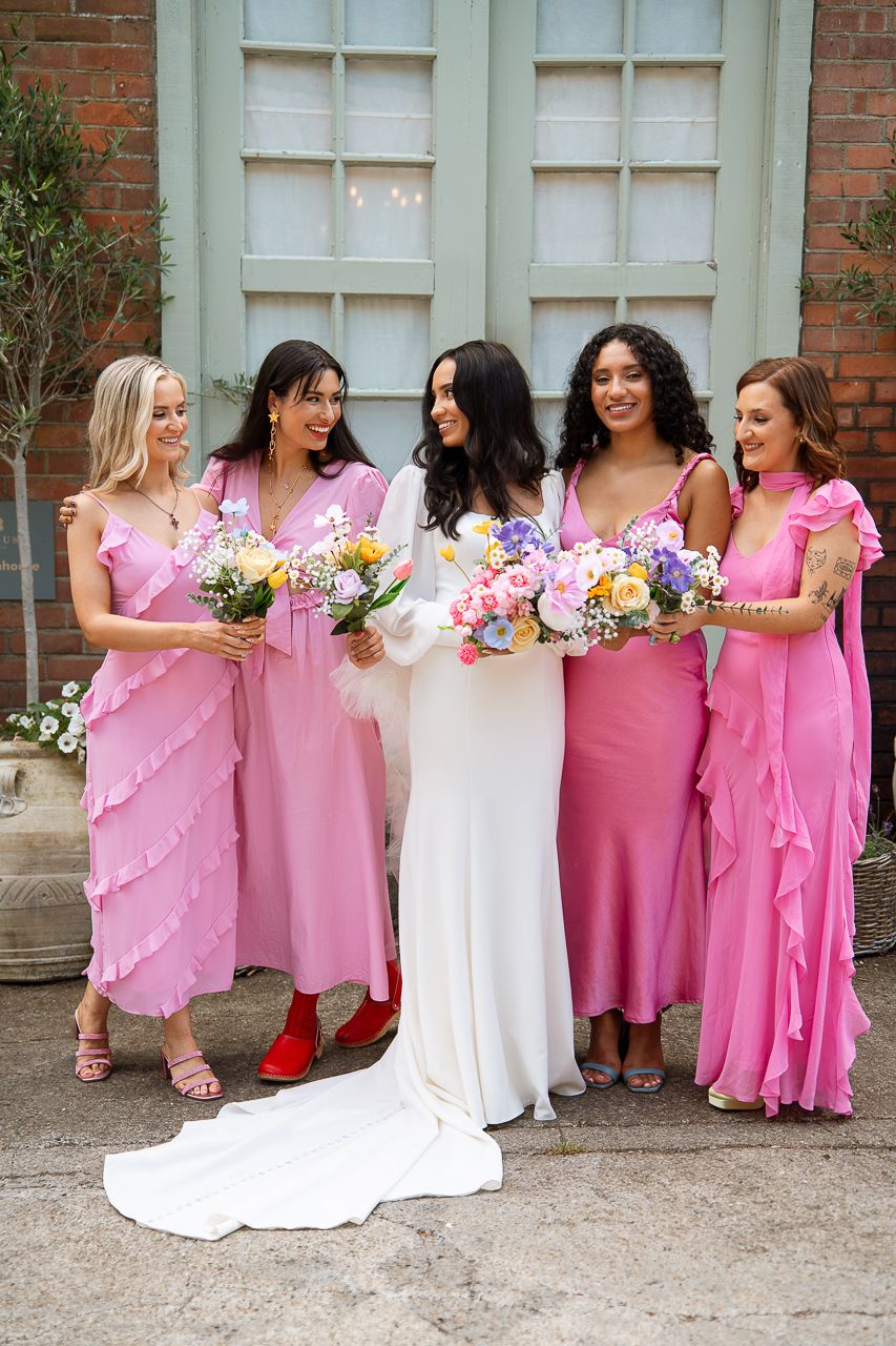 Wedding party in pink dresses and white gown gathered outside brick building with floral bouquets.