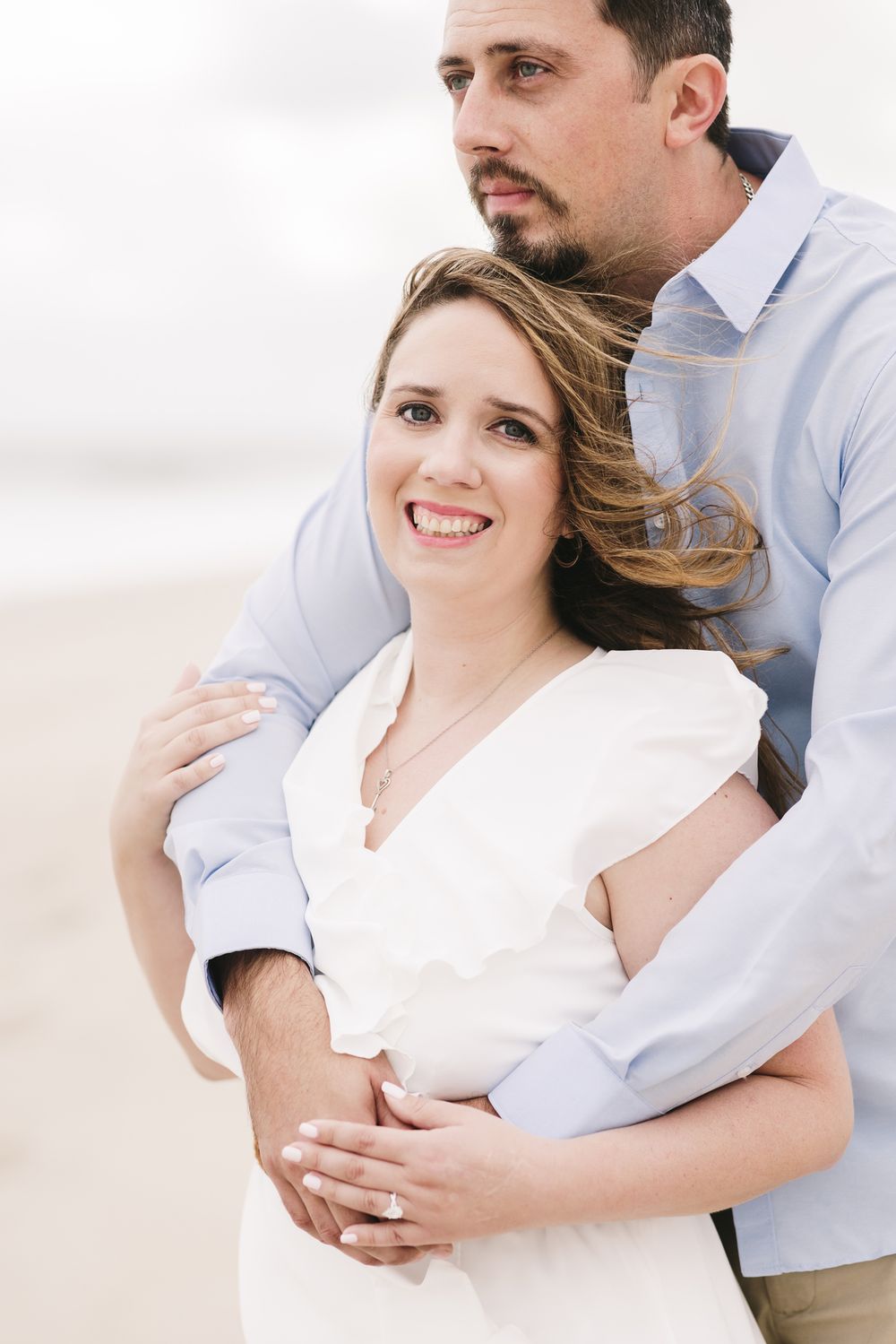 Woman in white smiles and embraced by fiancee in blue shirt looking away for engagement photos on cloudy beach day. Rye couple photographer