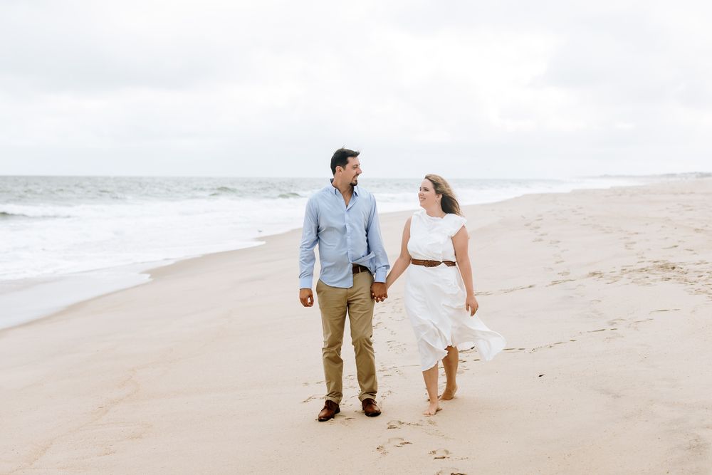 Couple holds hands walking on cloudy beach as breeze blows her white dress for engagement photos. North Shore MA engagement photographer.