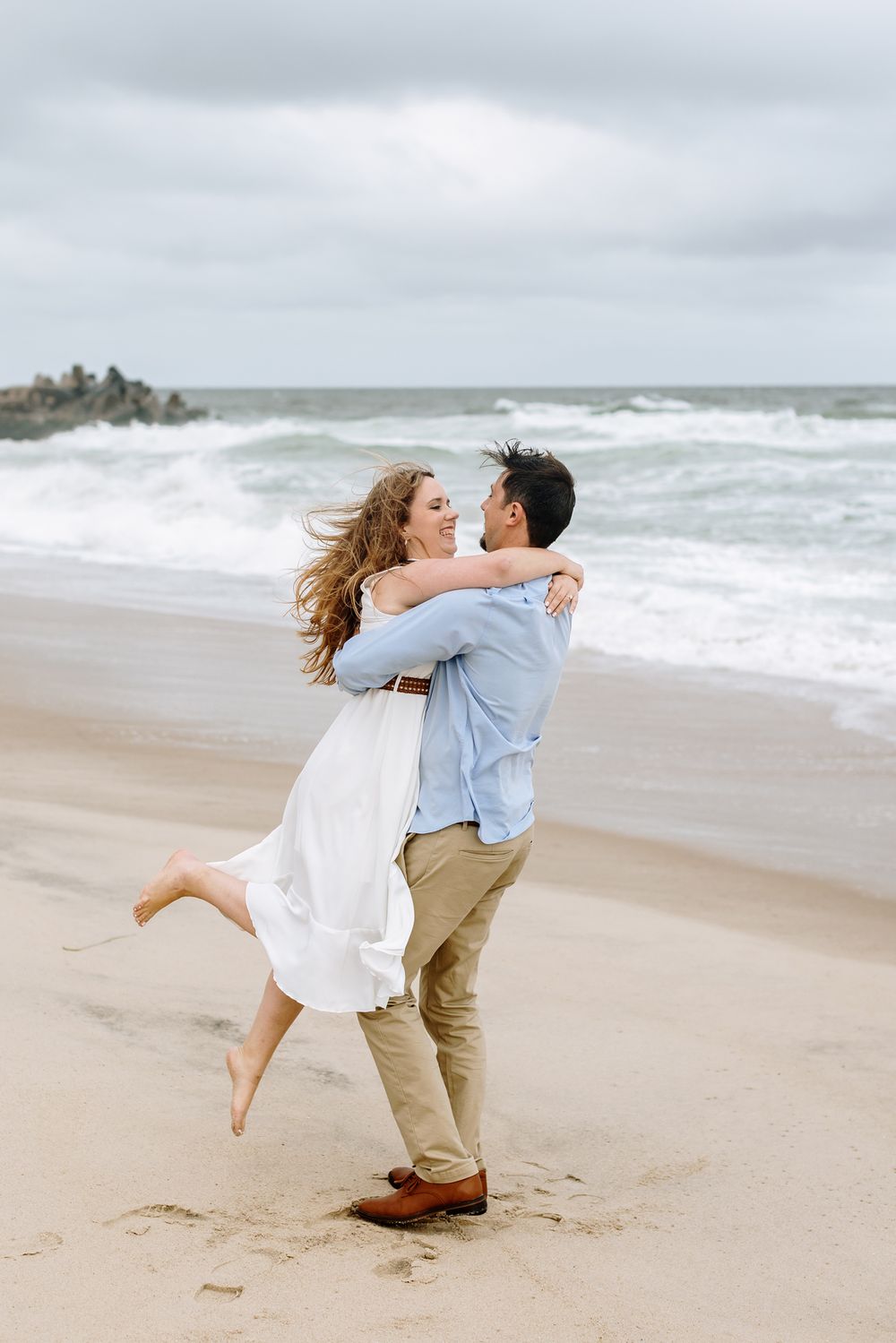 Woman in white dress is spun around by fiancee in blue shirt on cloudy beach day for engagement photos. Concord engagement photographer