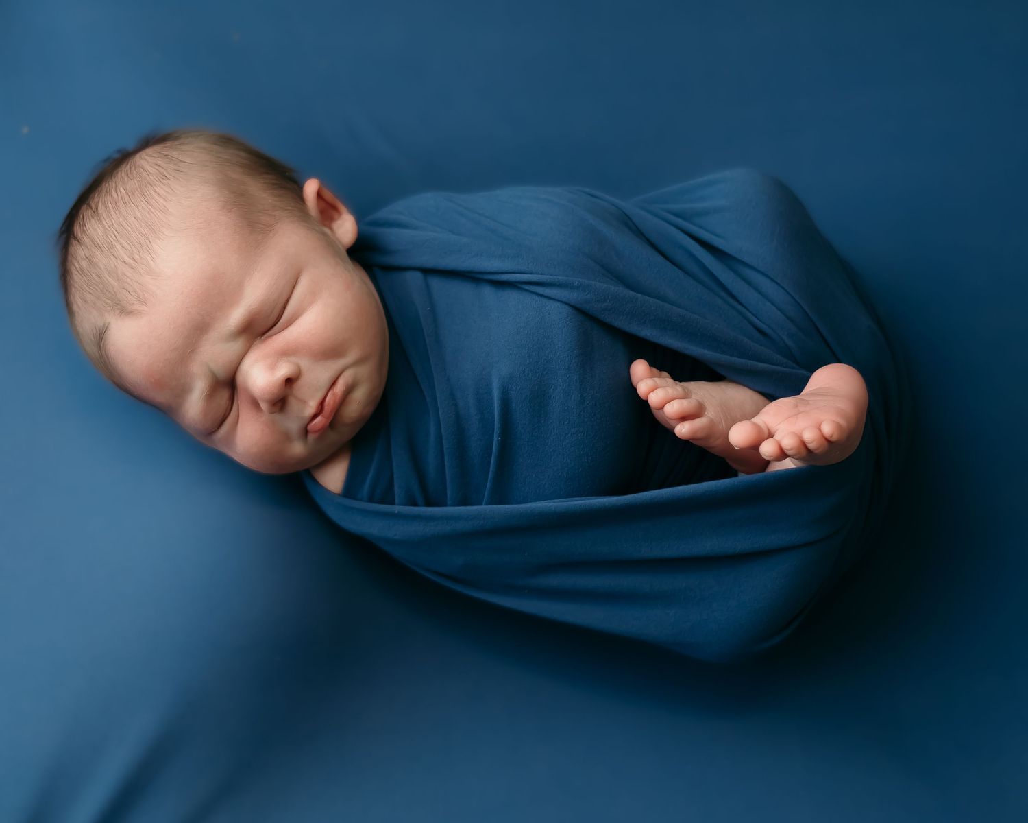 Sleeping newborn wrapped in soft blue fabric against blue background during professional photo session.