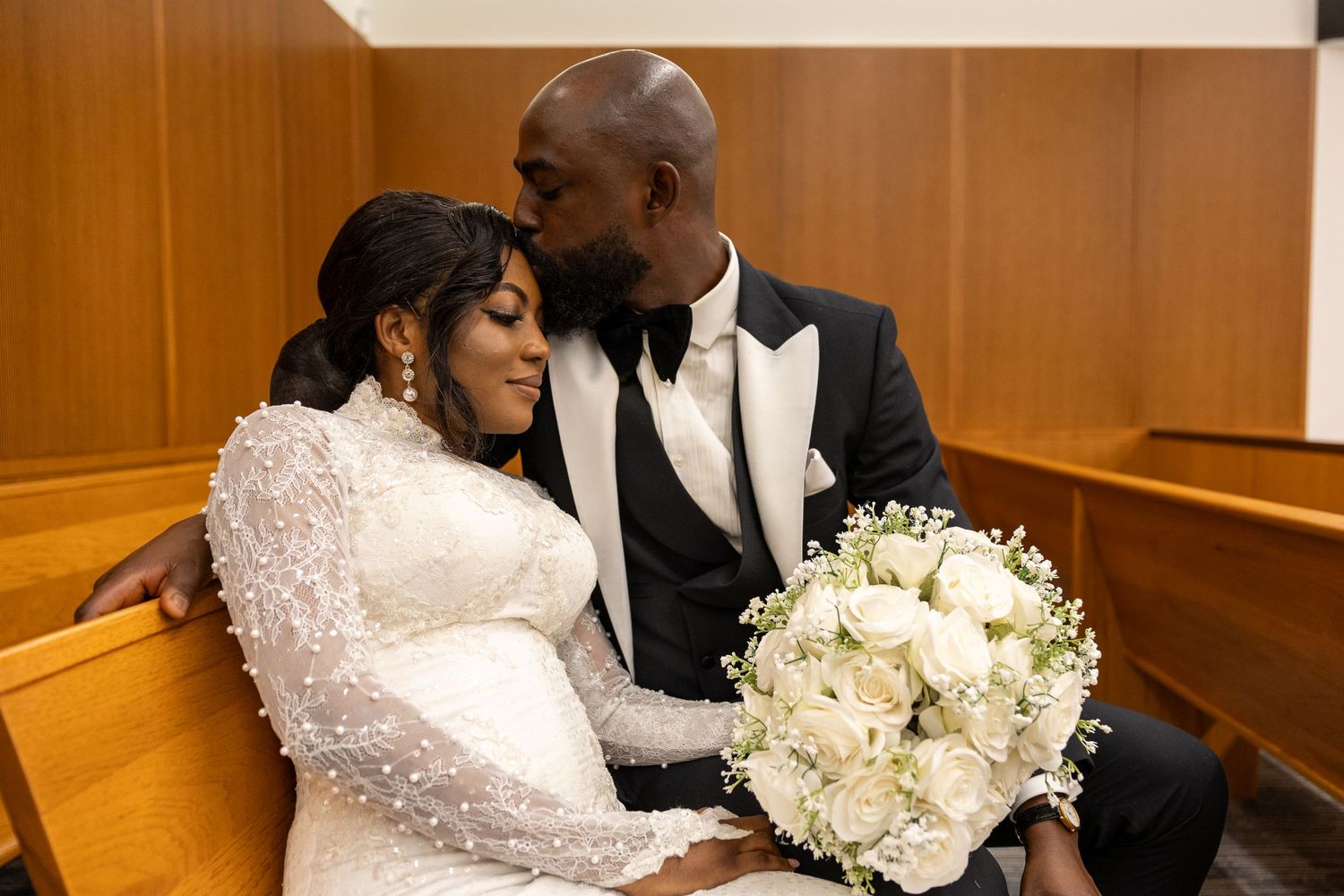 A bride in a white lace dress holds a white floral bouquet while sitting with her groom in formalwear.