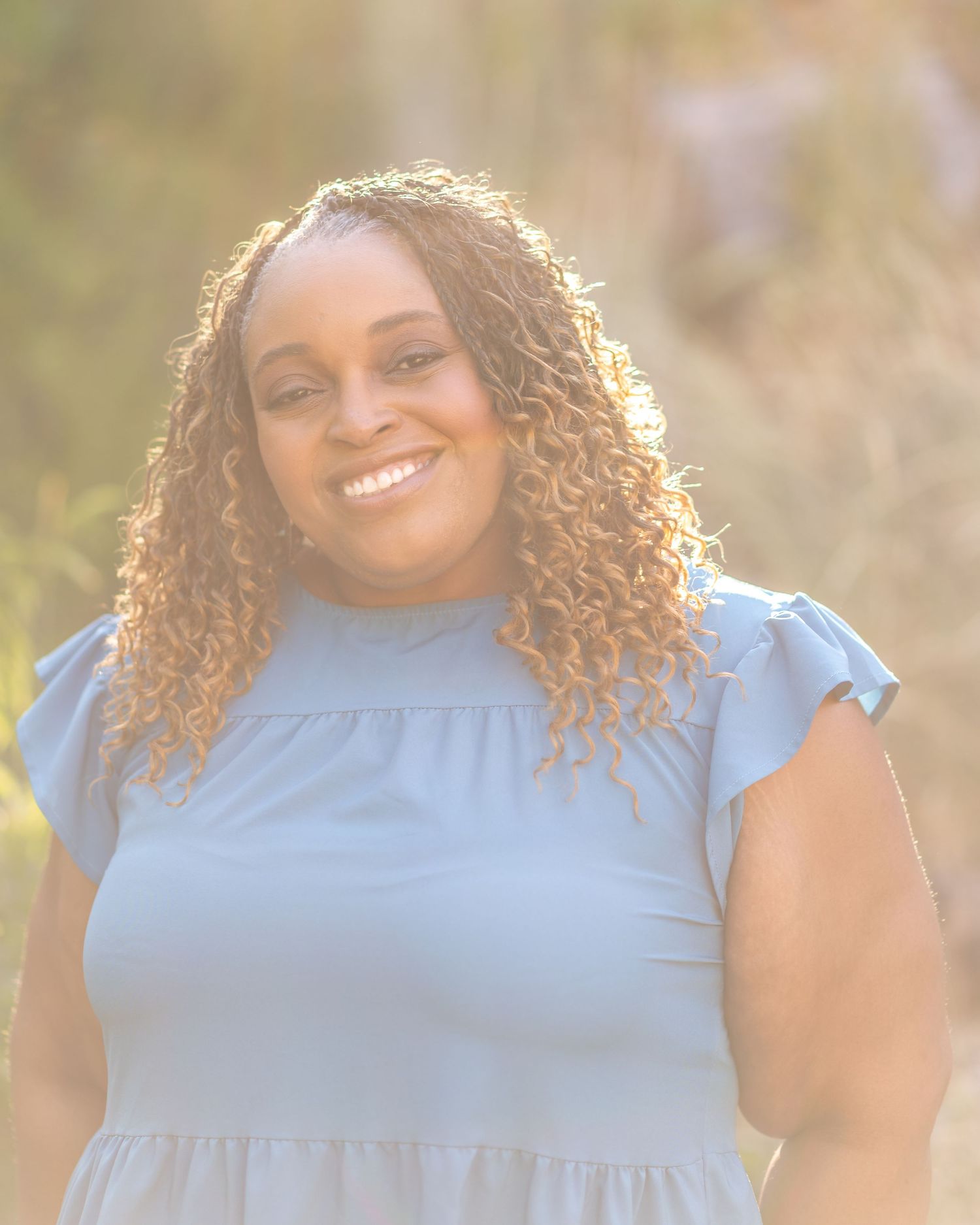 Outdoor portrait of a woman in blue dress with curly hair backlit by golden sunlight.