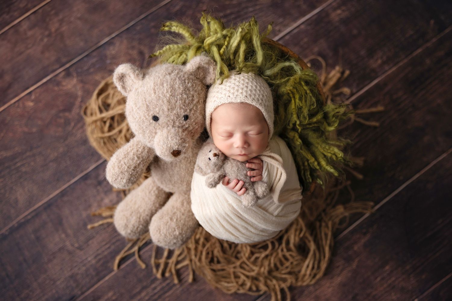 Sleeping newborn in white wrap next to plush teddy bear on wooden floor with greenery.