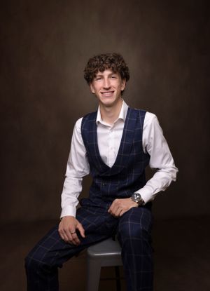 Young man in navy plaid vest and white shirt sitting against dark background.