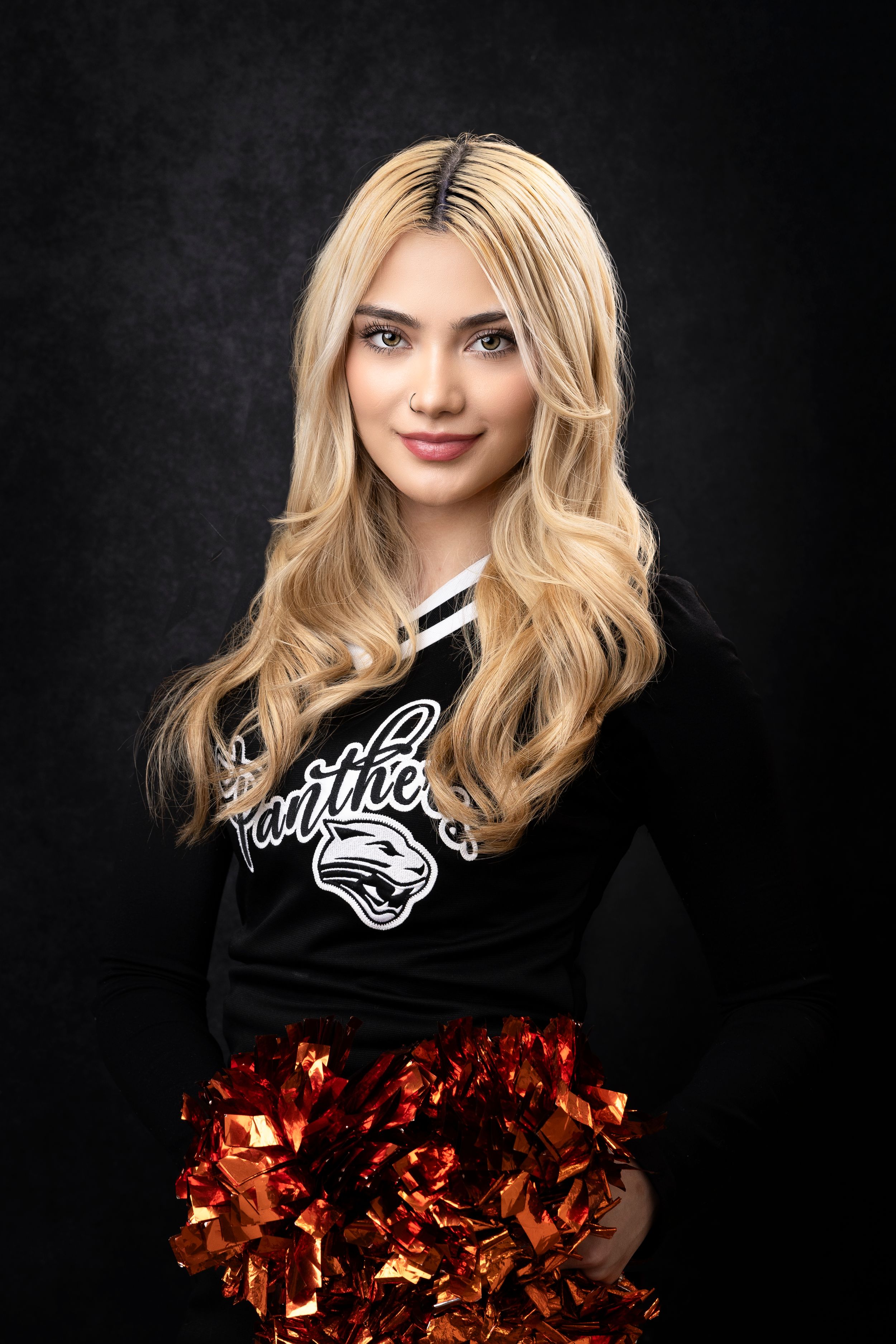Cheerleader in black uniform holding orange pom poms against dark backdrop.
