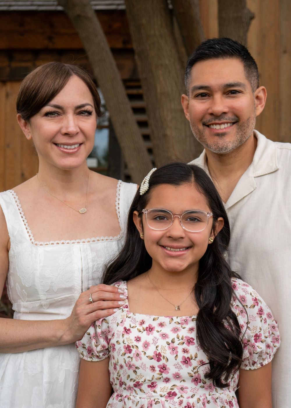 A smiling family of three poses outdoors, featuring a woman, man, and young girl in floral dress wearing glasses.