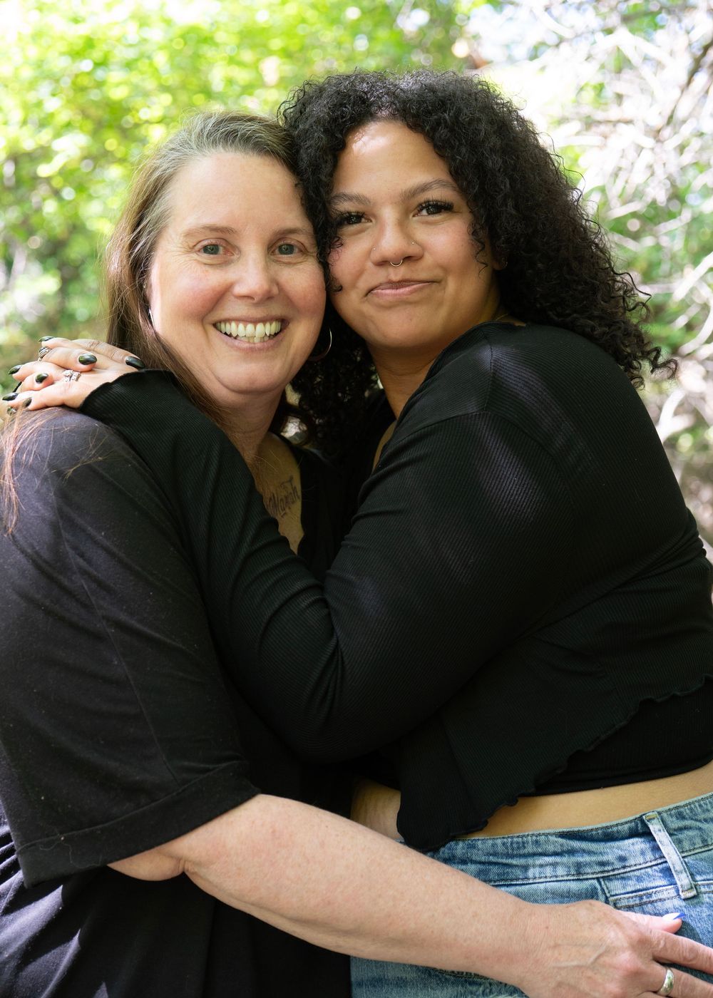 Two women hugging and smiling outdoors, sharing a warm embrace among green trees in natural light.