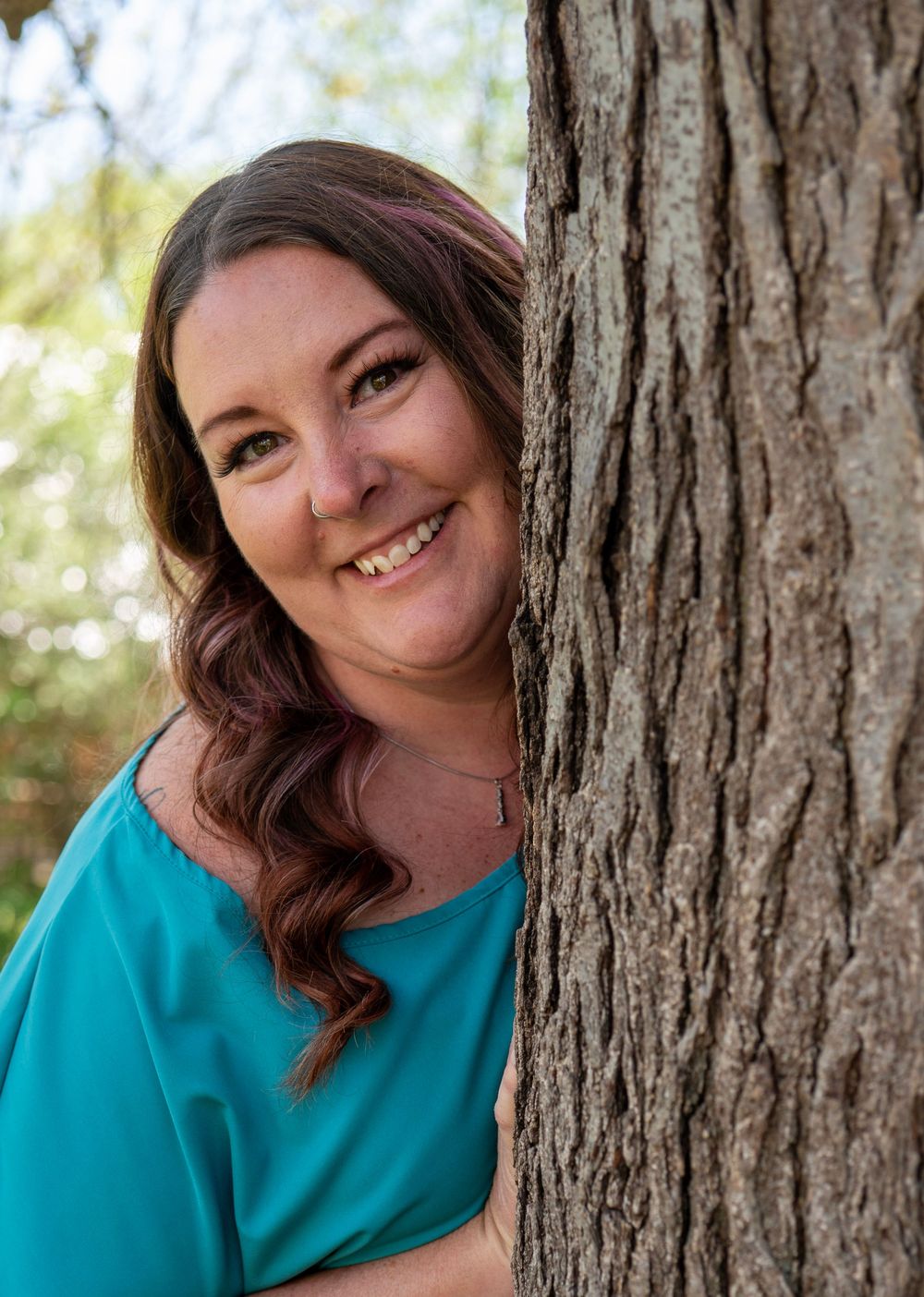 Smiling woman with auburn hair peeks around a tree trunk outdoors, wearing a teal top in a natural setting.