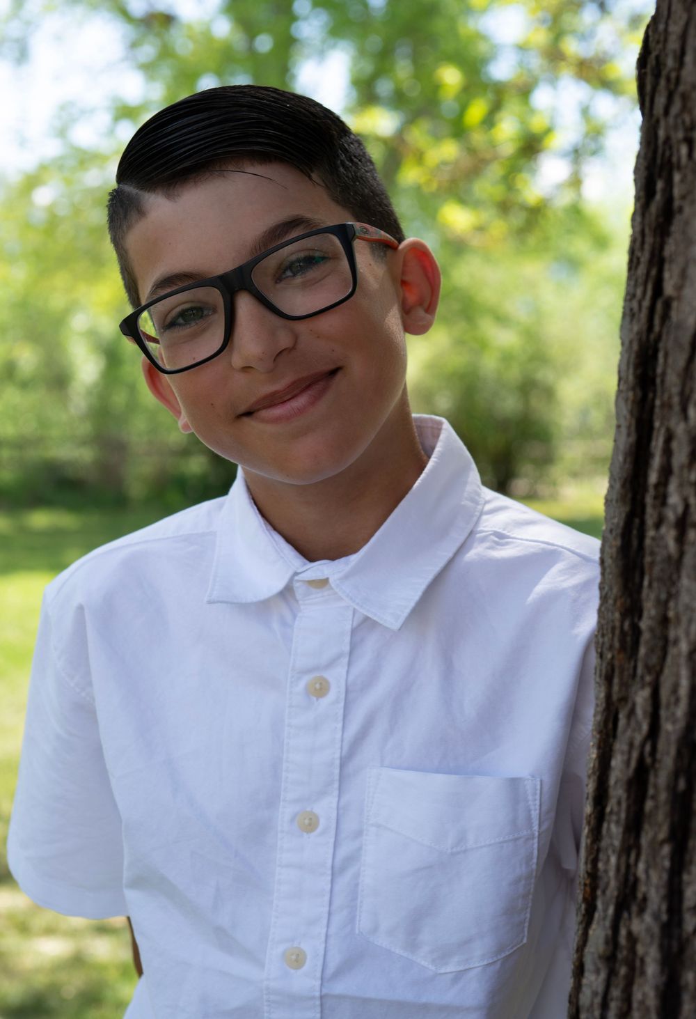 Smiling young boy with glasses in white shirt leaning against tree in outdoor park setting.