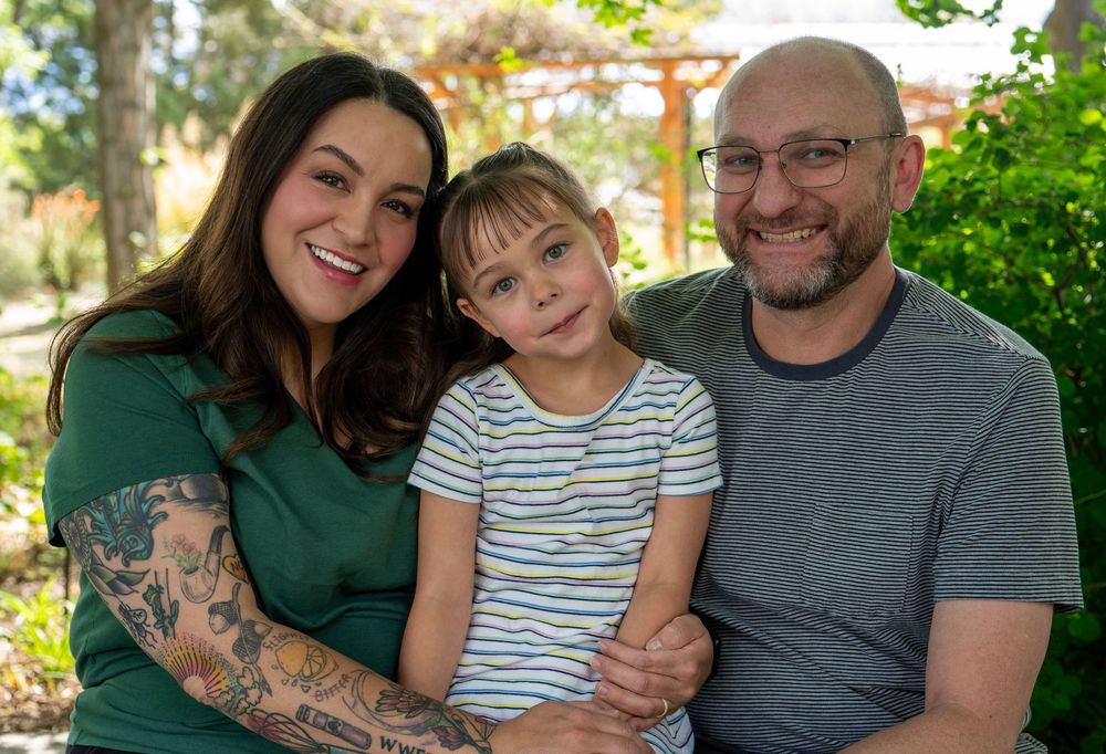 A smiling family of three poses outdoors, featuring a tattooed woman, young girl, and bearded man wearing glasses.