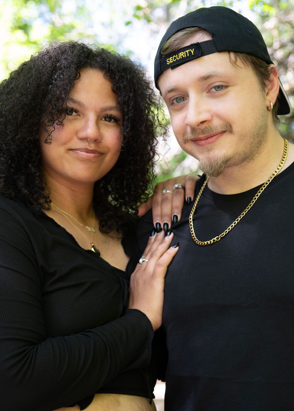 A young couple in black outfits poses together outdoors, the man wearing a black cap and gold chain necklace.
