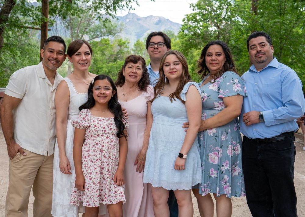 A joyful family of eight poses outdoors in pastel outfits with mountain trees in the background.