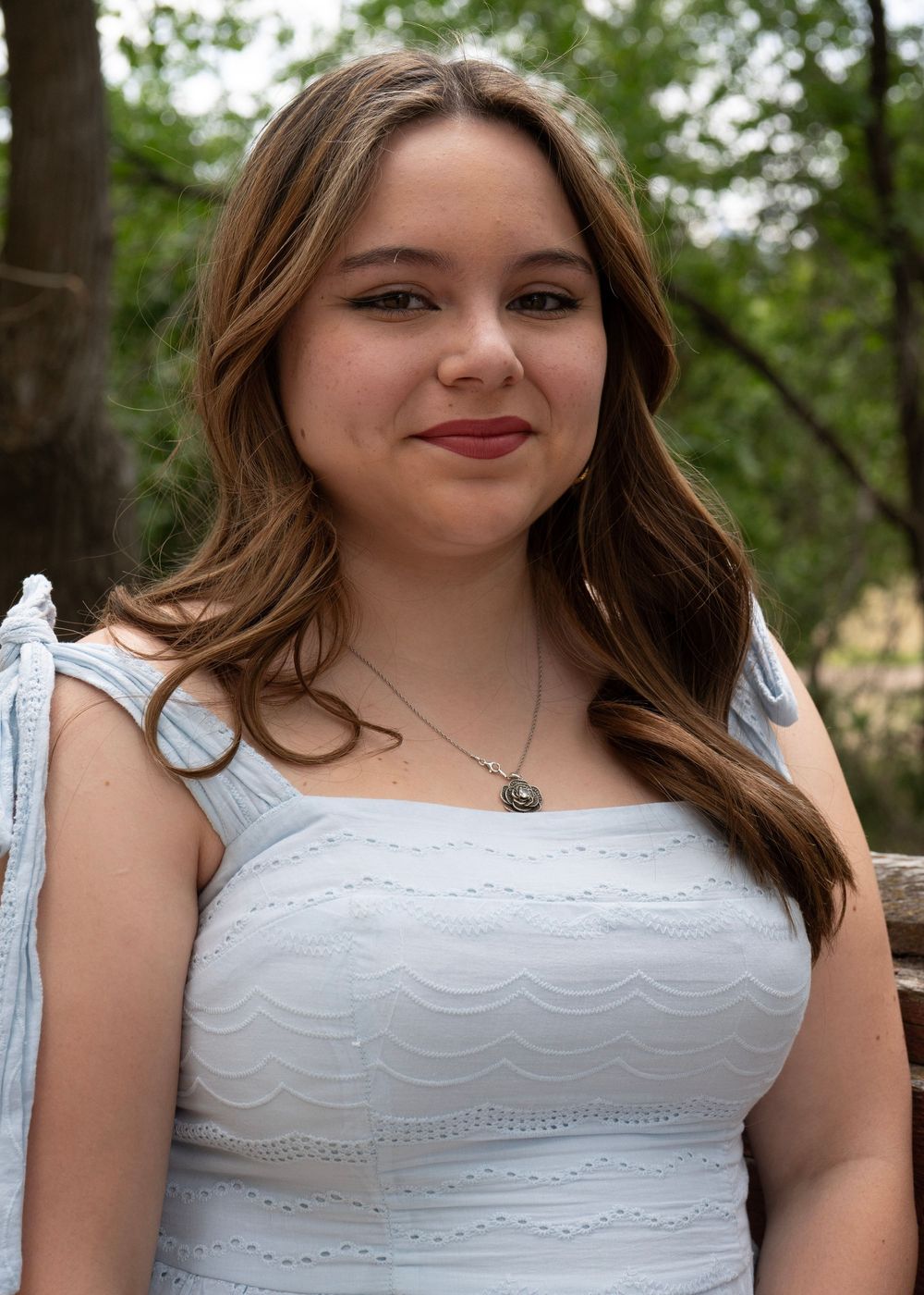 Young woman in white dress with necklace smiling outdoors among green trees.