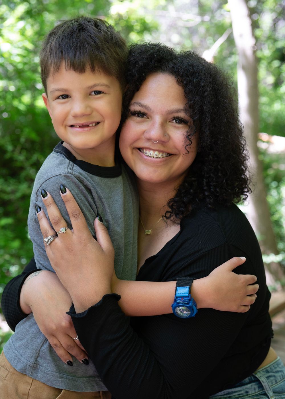 A woman with curly hair hugs a smiling young boy outdoors, both wearing casual clothing in a wooded setting.