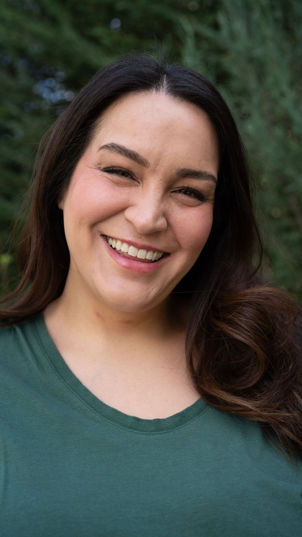Smiling woman with long brown hair wearing a green t-shirt outdoors with trees in background.