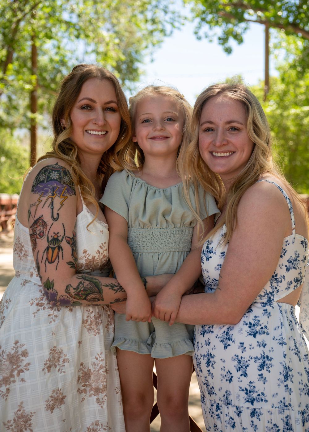 Two women in floral dresses smile with a young blonde girl outdoors among green trees on a sunny day.