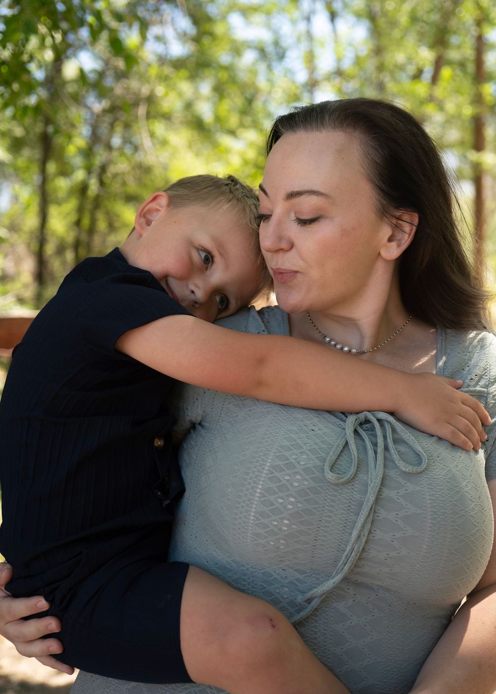 A woman with dark hair holds a young boy who hugs her neck outdoors among green trees.