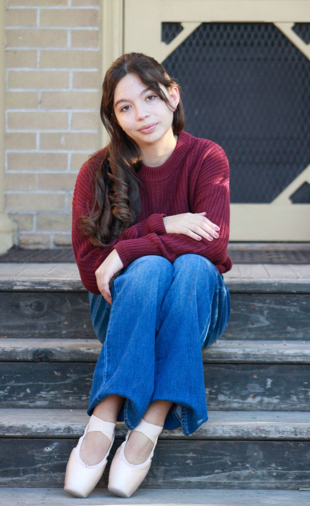 Person in burgundy sweater and jeans sits on outdoor steps near brick wall.