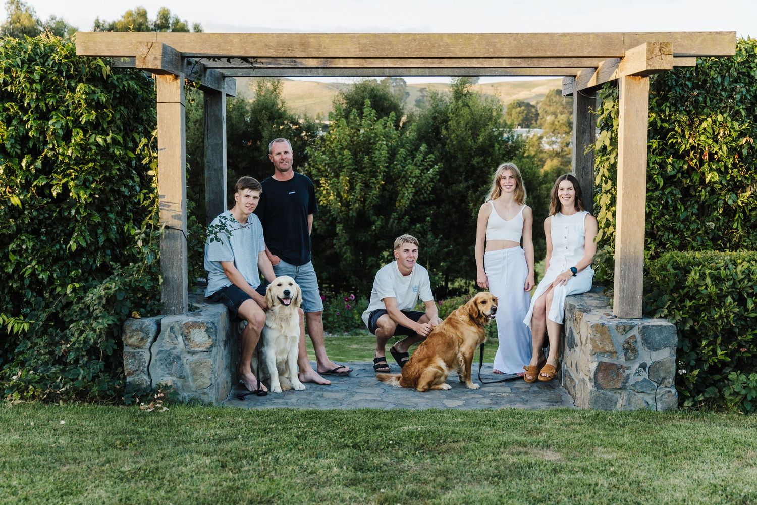 Family portrait under a garden pergola with two dogs and lush greenery.
