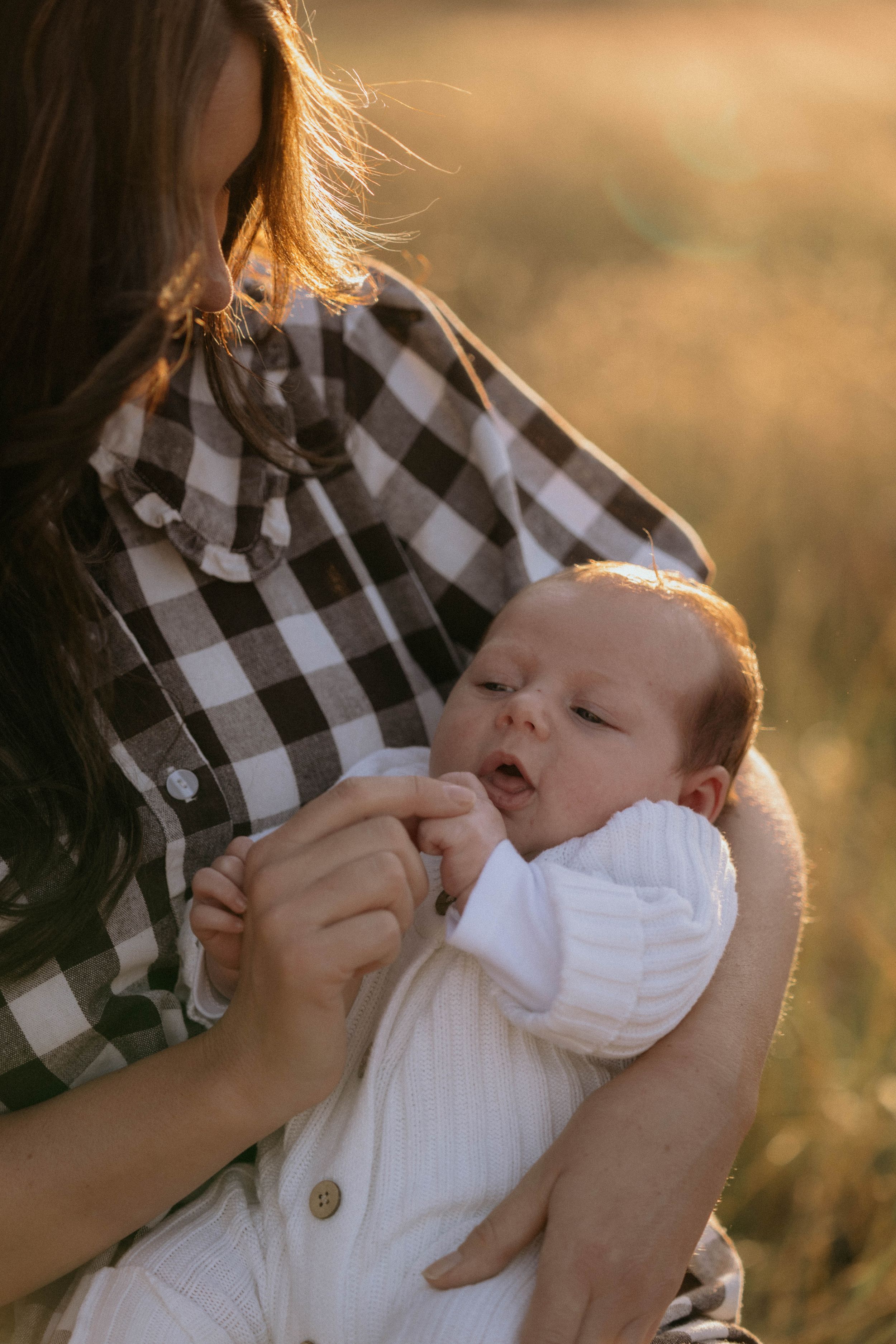 Mother in buffalo plaid shirt holding small baby in white outfit during golden hour.