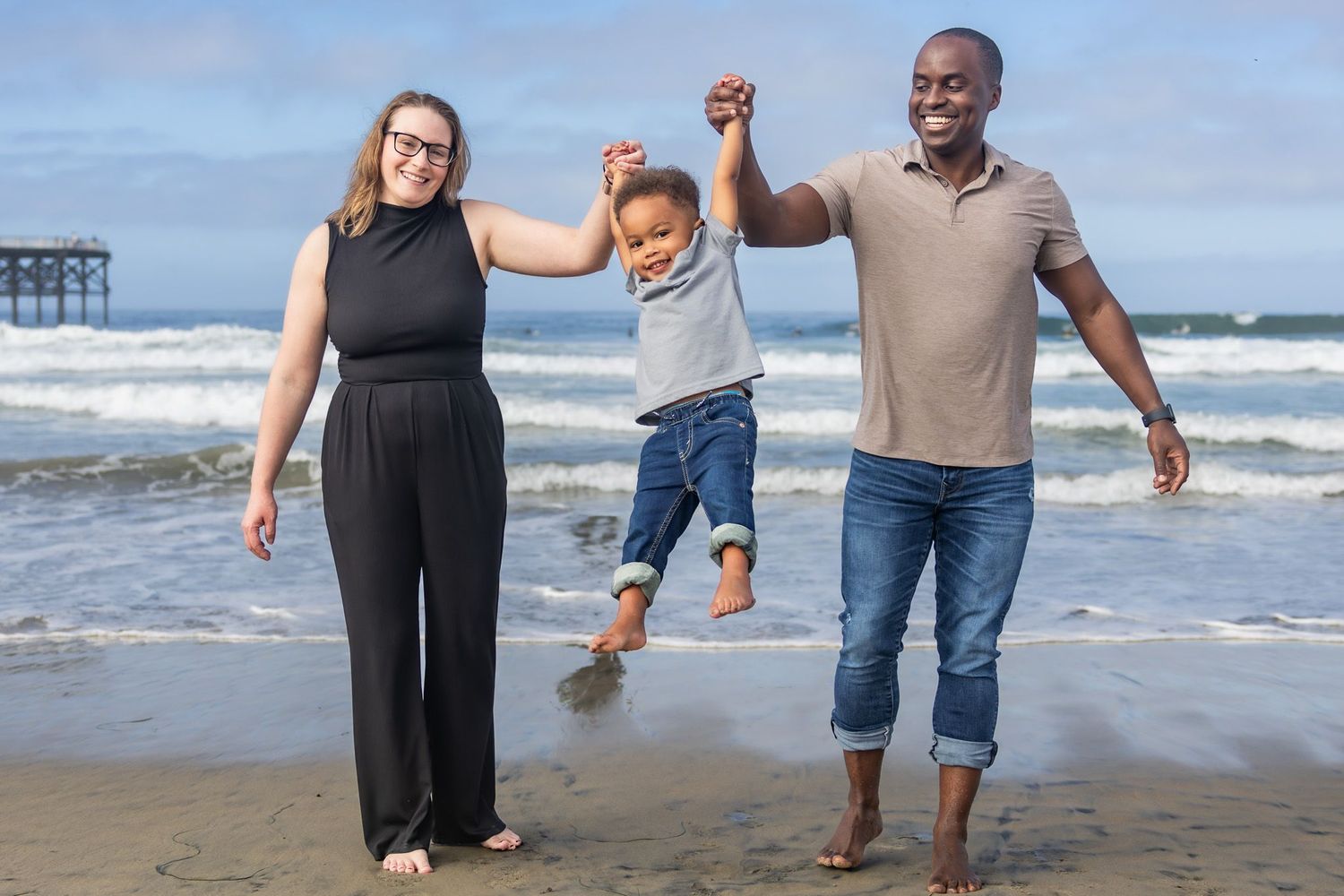 San Diego family portrait photography of parents swinging their young child on a beach with ocean waves and a pier in the background.