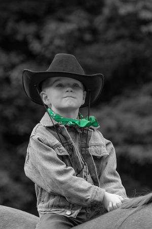Black and white photo of a rider wearing a cowboy hat, denim jacket and bright green bandana while horseback riding.