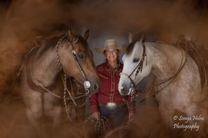 A person wearing a cowboy hat kneels alongside three horses in a shadowy rustic setting.