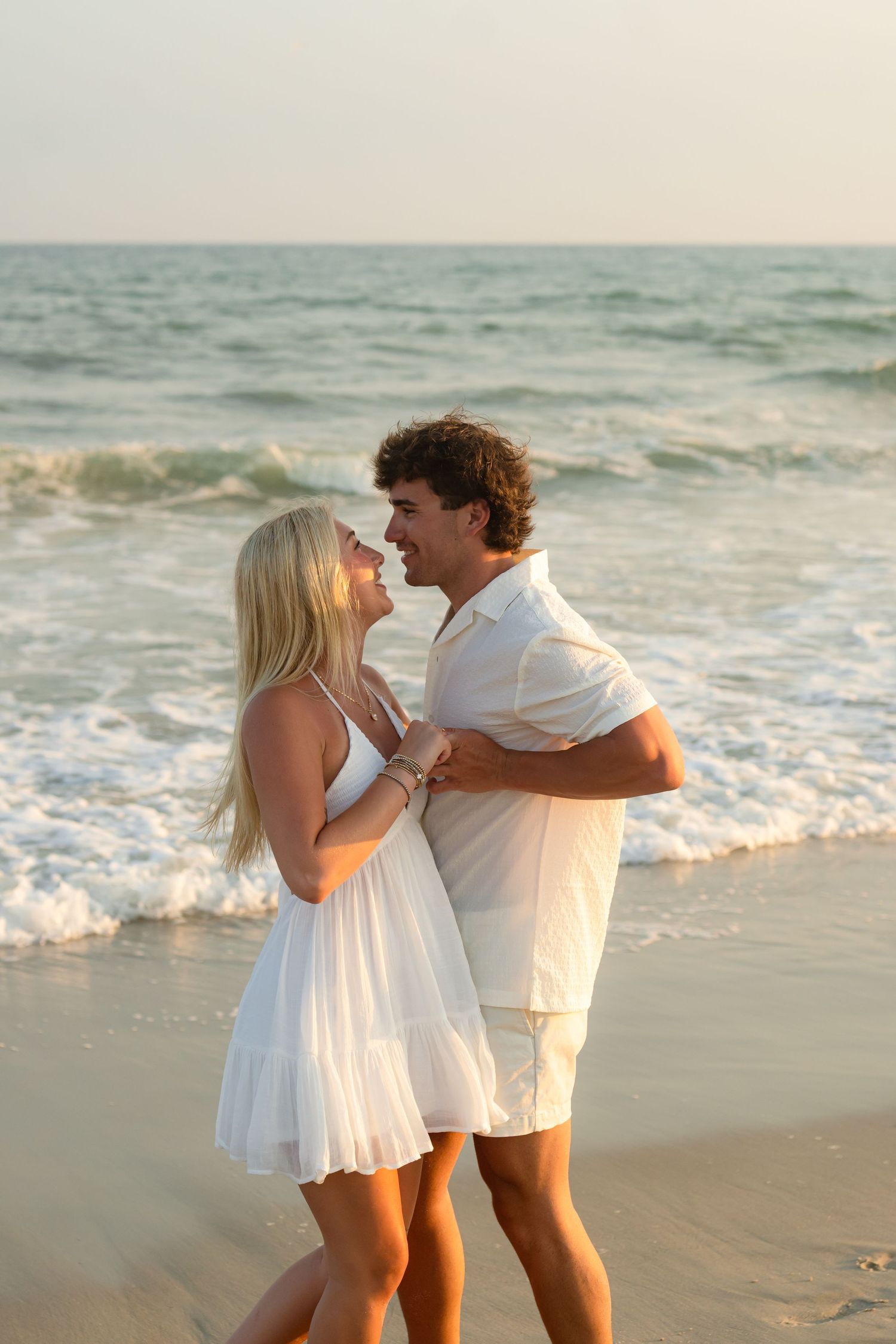A romantic couple embraces under a pier by the ocean at sunset.