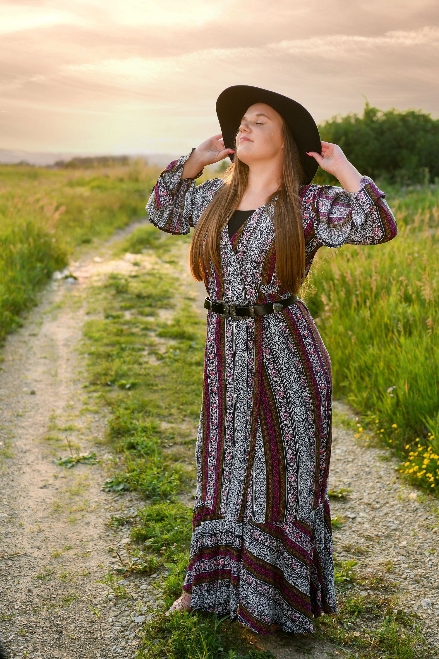 A long-haired figure in a flowing bohemian maxi dress and hat poses on a dirt path at sunset in a grassy meadow.