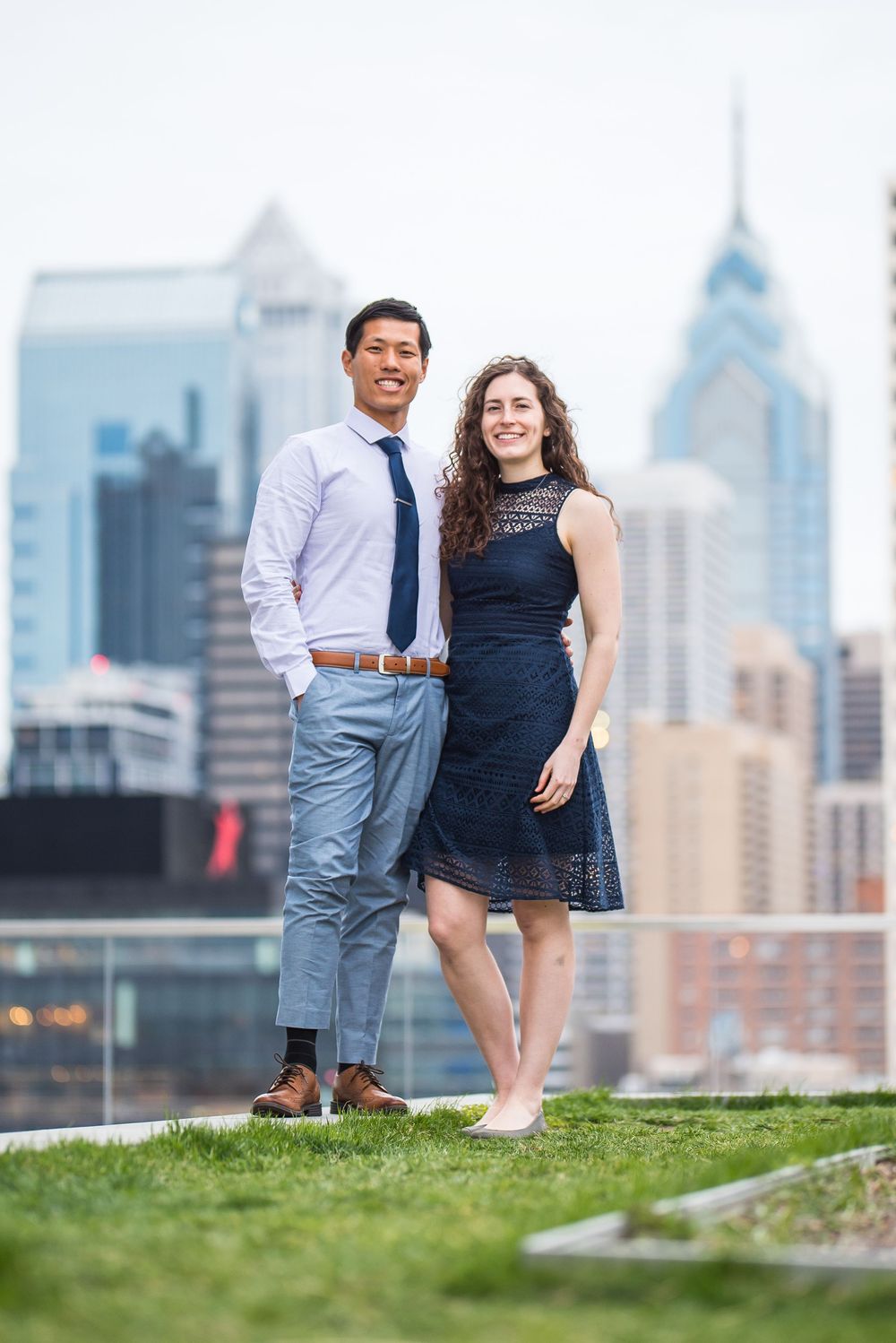 A couple poses for engagement photos on a grassy hill overlooking the Philadelphia skyline on a bright day.