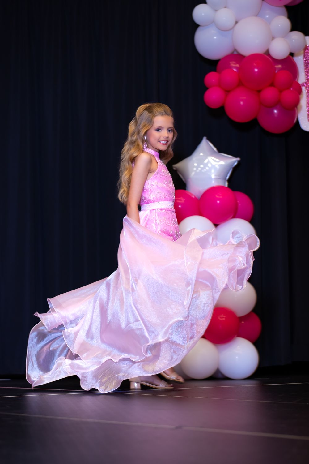 Young performer in pink gown twirls on stage with balloon decorations.