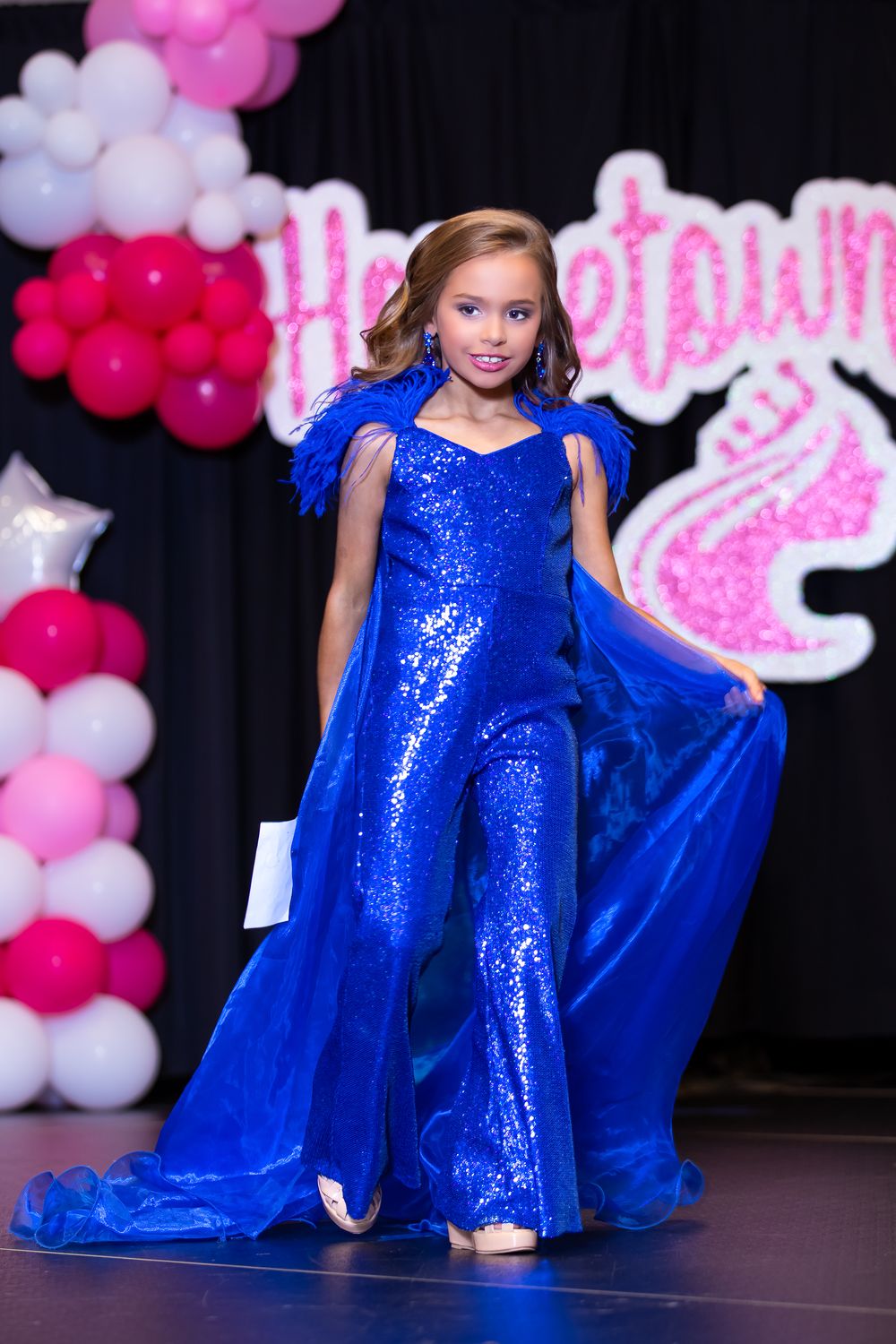 Stage performer in royal blue sequined jumpsuit poses with flowing cape detail.