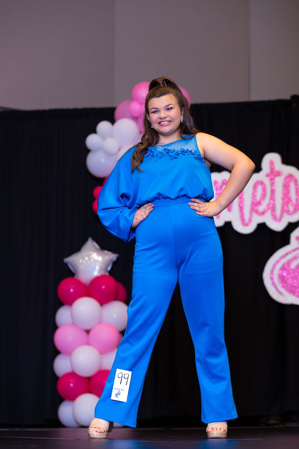 Model in royal blue one-shoulder jumpsuit poses confidently on stage.