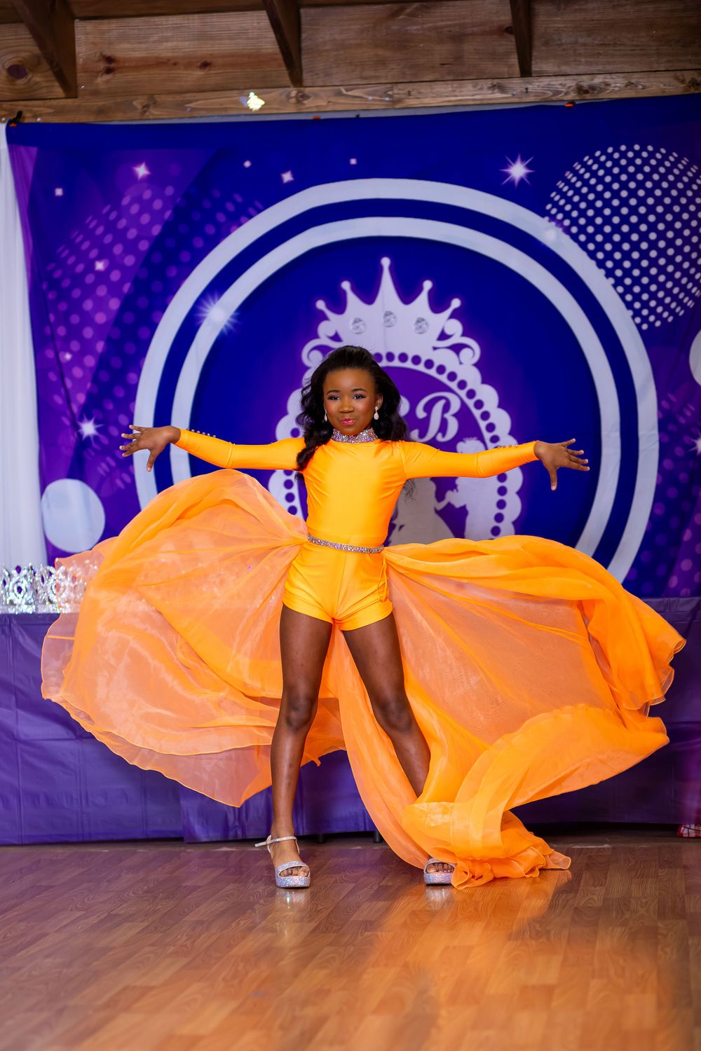 Energetic dance pose with arms spread wide in yellow costume with flowing orange fabric.